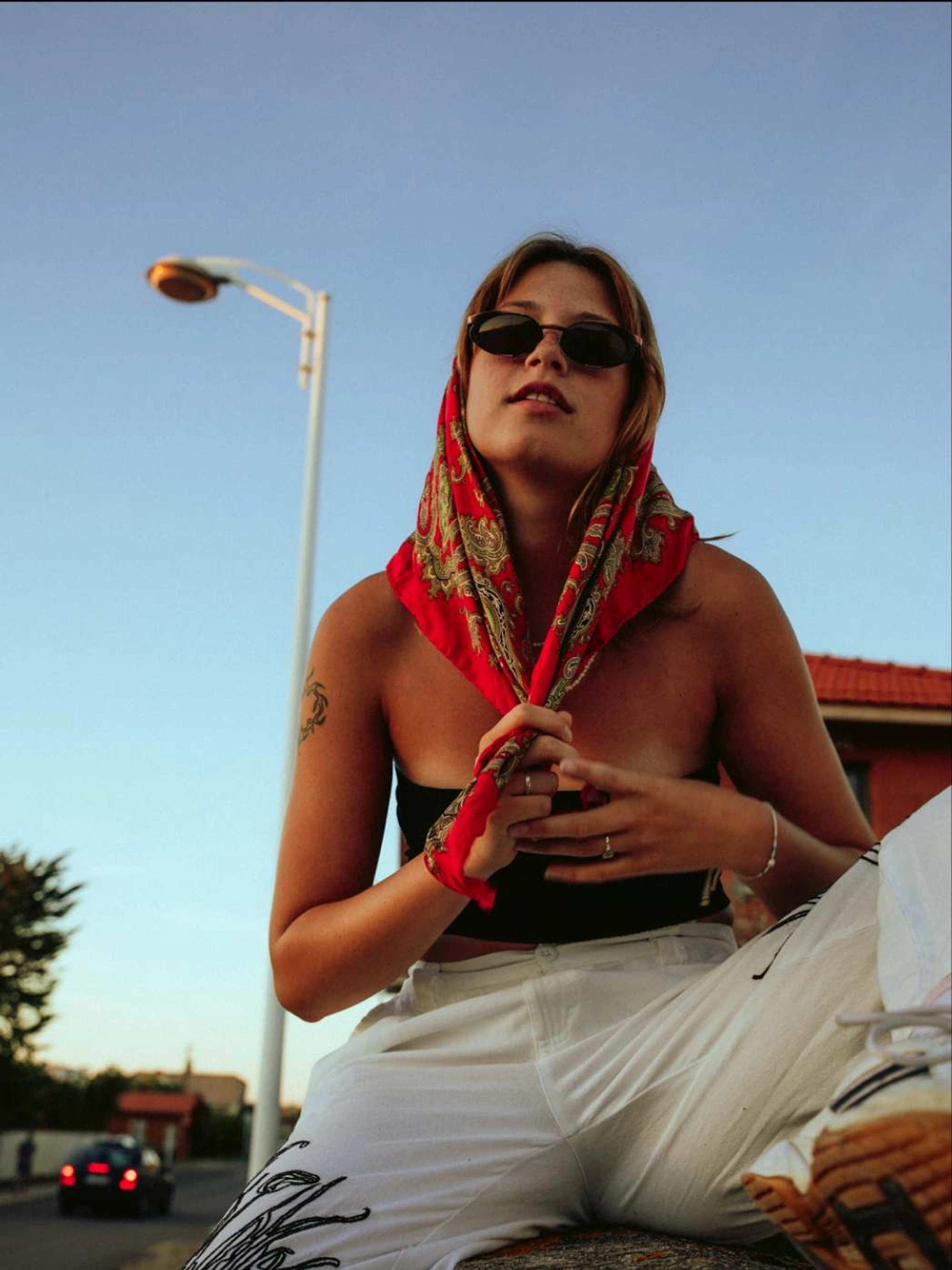young woman wearing sunglasses sitting on a wall, wrapping a scarf around her head